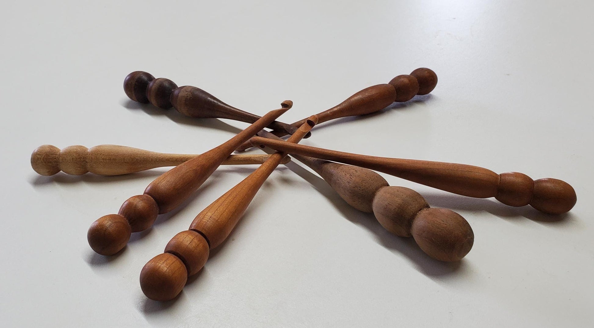 A small, artfully arranged pile of hand-turned 5mm crochet hooks made from maple, cherry, and walnut wood. Each hook is polished smooth and showcases unique natural grain patterns. The light maple, warm cherry, and dark walnut woods create a beautiful contrast in color and texture. The ergonomic handles and glossy finish reflect craftsmanship and care, with the hooks overlapping gently in a visually pleasing, organic composition.