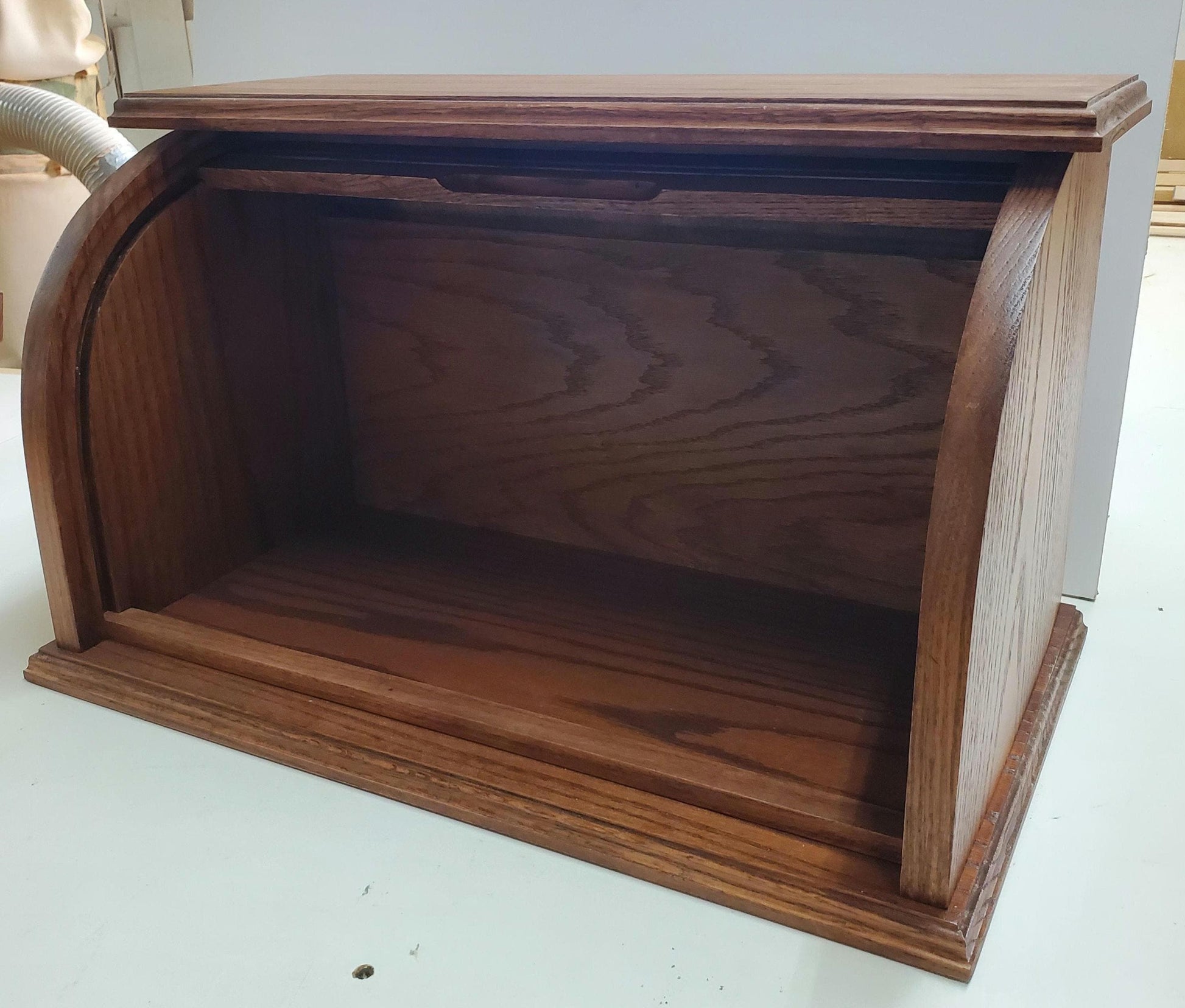 Oak bread box with a roll-top door, crafted from natural oak wood that beautifully highlights the grain. Stained in a medium shade of reddish-brown for a rich, inviting look, it offers a warm, rustic touch to any kitchen. The spacious interior helps keep bread and baked goods fresh, while the sturdy construction and classic design add both style and convenience. Shown from the front with the door fully open, showcasing its elegant design and practical functionality.