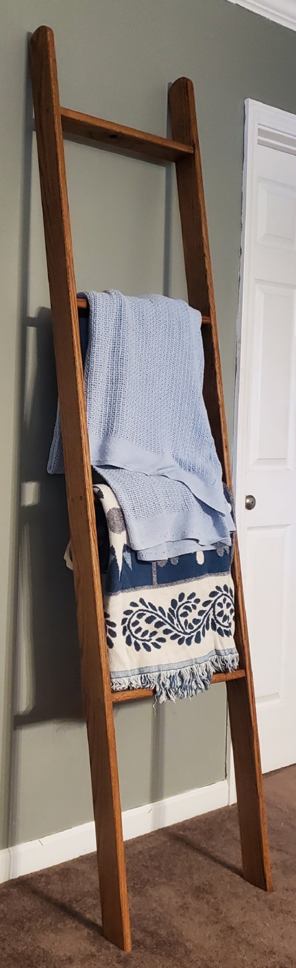 Wooden oak blanket ladder in a rich Early American stain, leaning against a plain wall with soft blue blankets hanging on it. The warm, rustic finish highlights the natural oak grain, adding a cozy, library-inspired charm to any space. Perfect for displaying blankets, throws, or towels, this sturdy ladder combines style, functionality, and vintage flair to enhance your home décor.