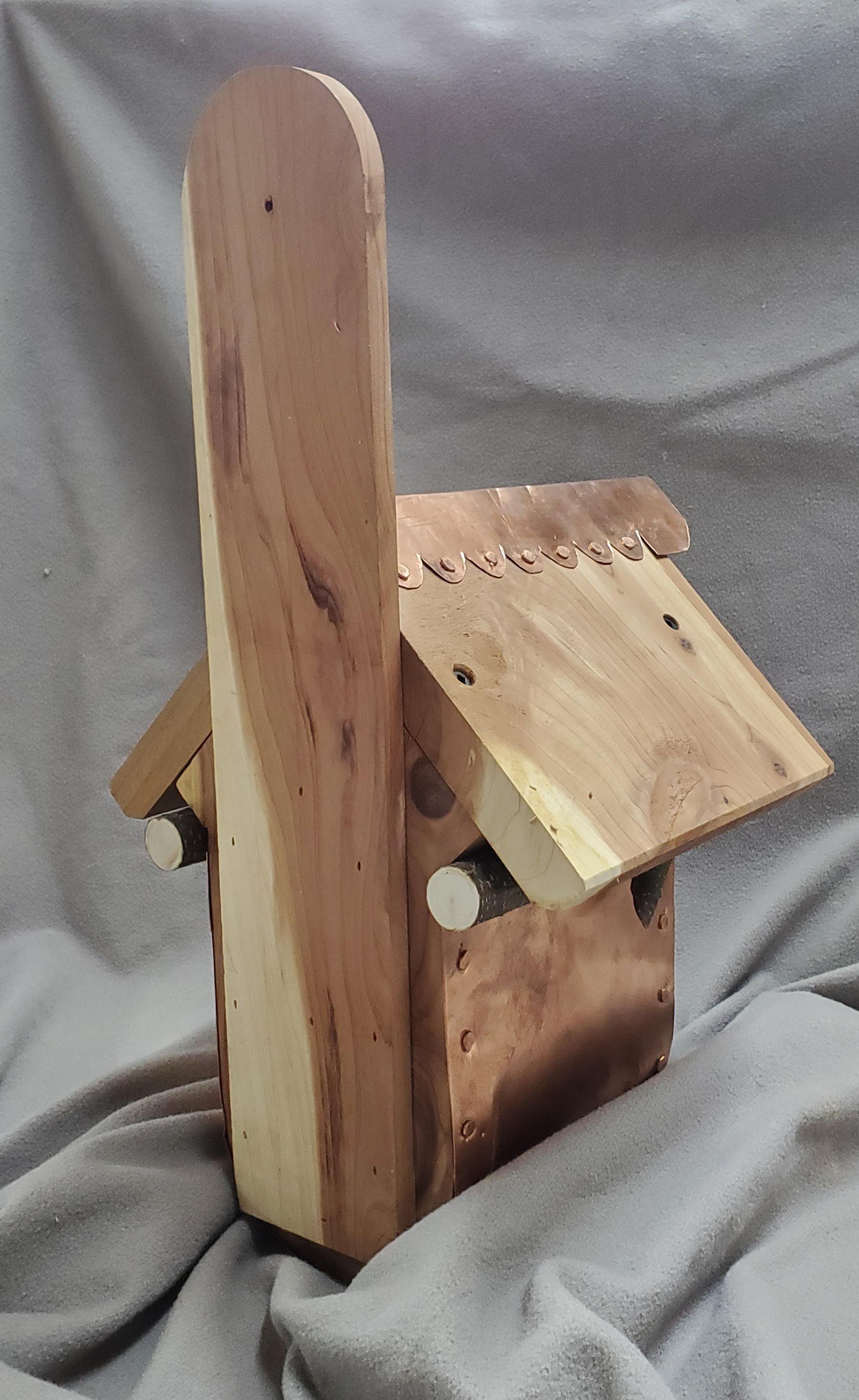 Back view of a handcrafted birdhouse made from warm-toned cedar wood with a curved copper bottom. The cedar roof slopes gently downward, showcasing the natural grain and texture of the wood. A copper ridge cap runs along the peak of the roof, providing both decorative detail and weather protection. The combination of polished copper and rustic cedar highlights the artisanal craftsmanship of this durable outdoor birdhouse.