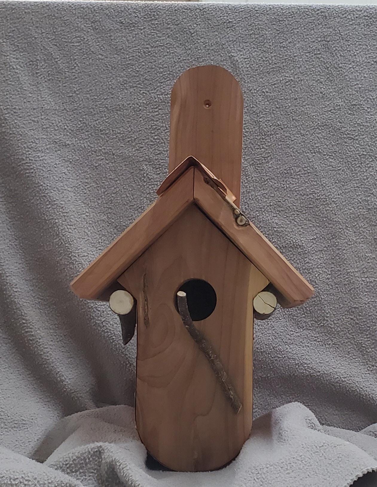 Front view of a handcrafted birdhouse featuring a curved copper bottom and a warm-toned cedar roof. A copper ridge cap runs along the roof peak, adding both decorative detail and weather protection. The entrance hole is centered on the front face of the birdhouse, framed by the natural grain of the cedar wood. The mix of polished copper and rustic wood creates a striking contrast, highlighting the craftsmanship and charm of this unique outdoor piece.