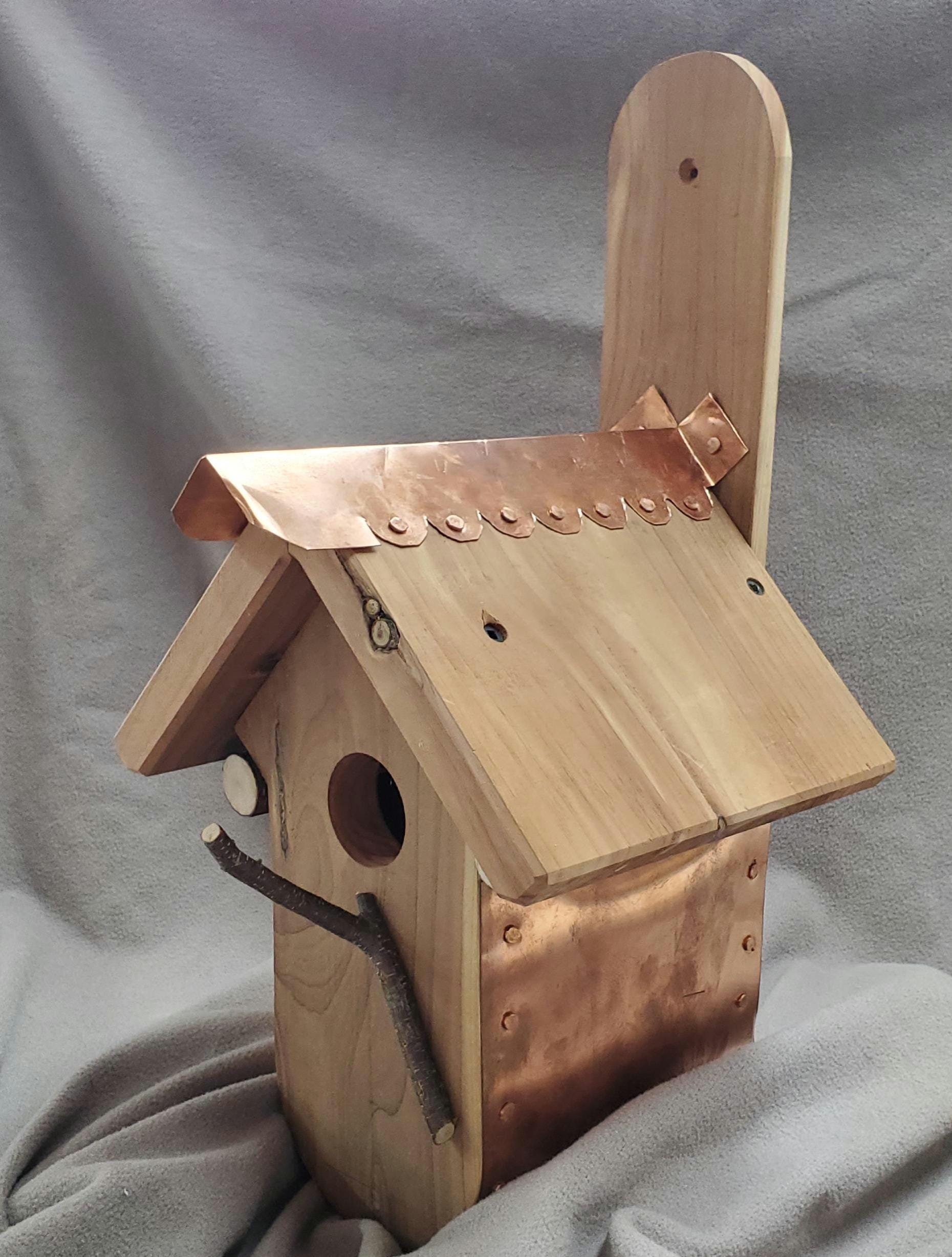 Angled side view of a handcrafted birdhouse designed for bluebirds, featuring a curved copper bottom and a warm cedar roof with a copper ridge cap along the peak for weather protection. The entrance hole is visible on the front panel, framed by the natural cedar grain. The polished copper contrasts beautifully with the rustic wood, highlighting the craftsmanship. This birdhouse blends functionality and aesthetic appeal in a unique outdoor design.