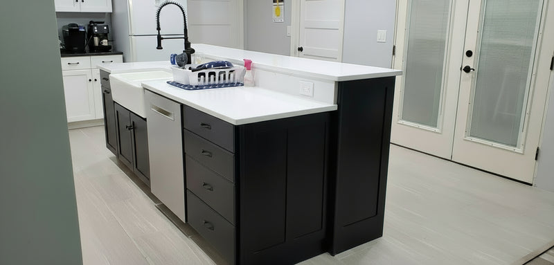 Modern kitchen with black tiered island and white countertops featuring a dishwasher , stack of drawers, a porcelain farmhouse sink, and a trash pullout